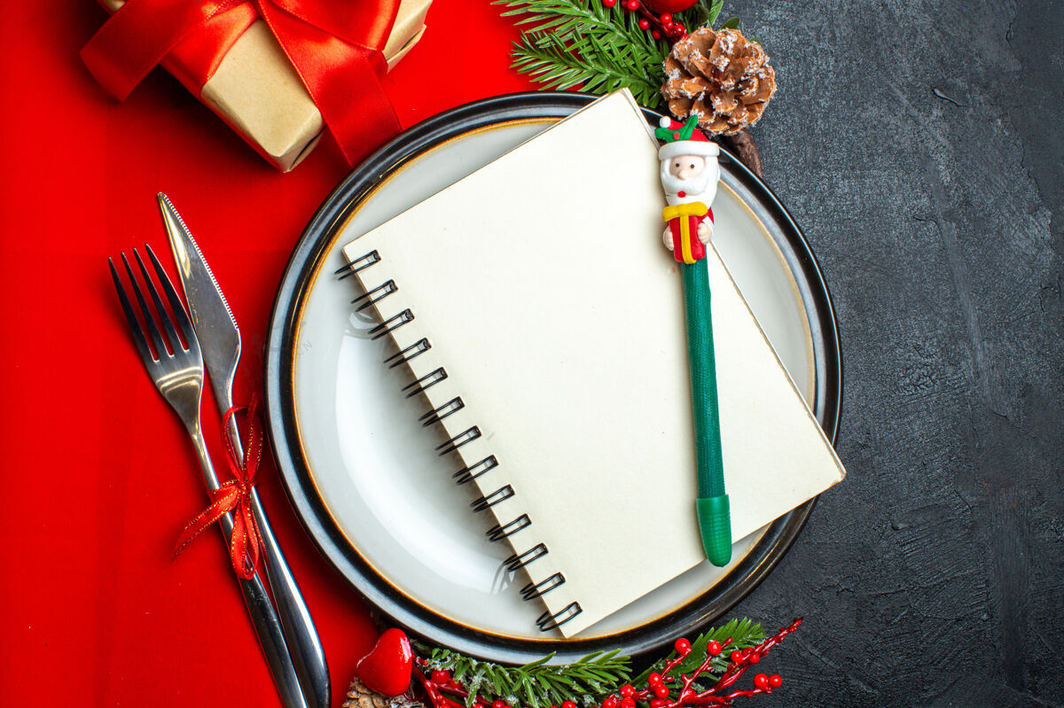 圣诞新年背景特写图 餐盘上有螺旋笔记本 餐具套装 装饰配件杉木树枝 旁边是一张黑色桌子上的红色餐巾上的礼物螺旋鼓树枝