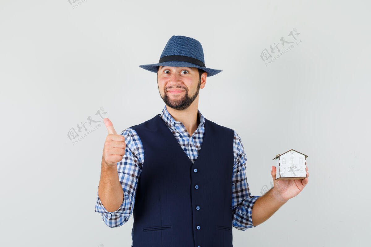 前面年轻人举着房子的模型 在衬衫 背心 帽子和看起来快乐 正面视图拇指向上男人模特休闲