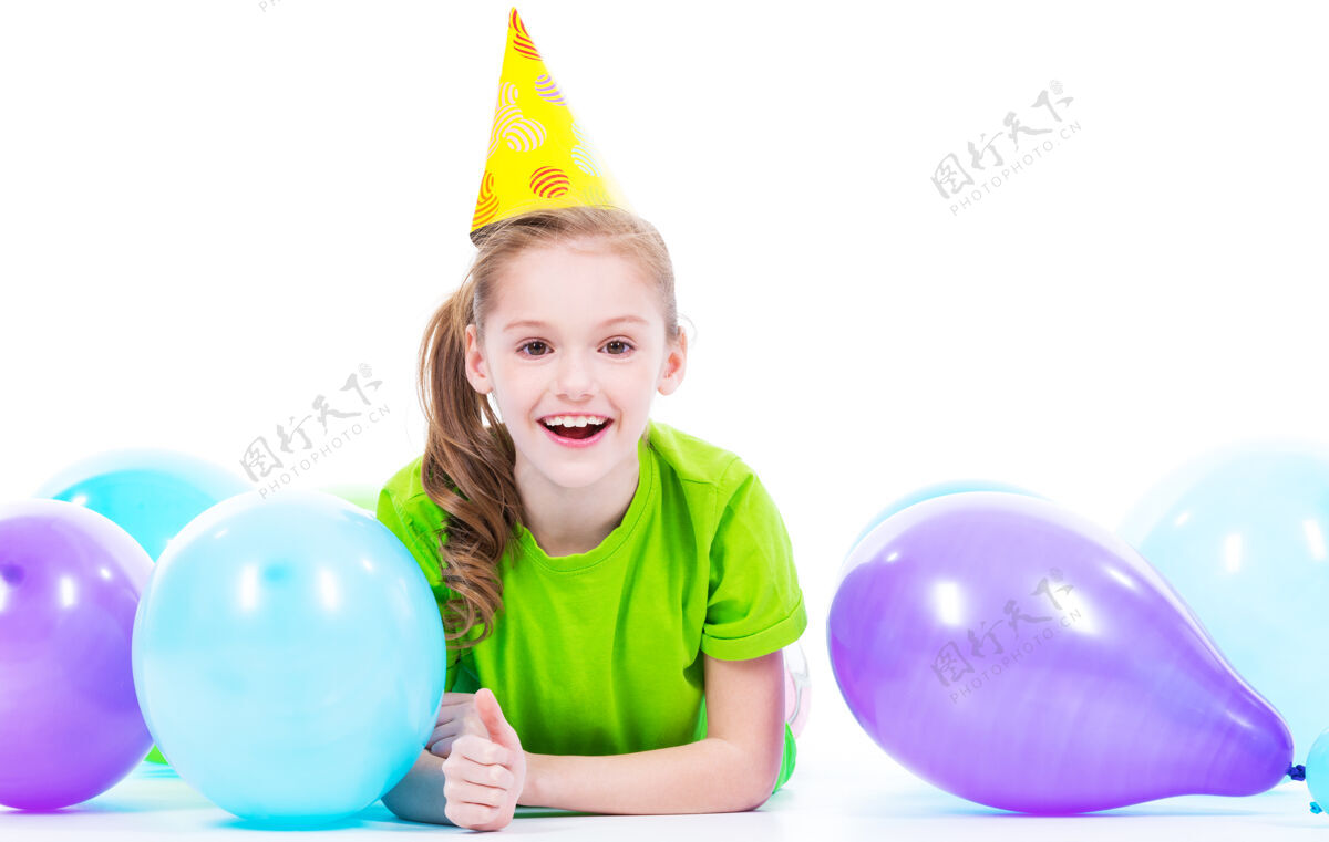 欢呼穿着绿色t恤的快乐微笑女孩躺在地板上 带着五颜六色的气球-隔离在白色的地板上竖起大拇指白种人庆祝