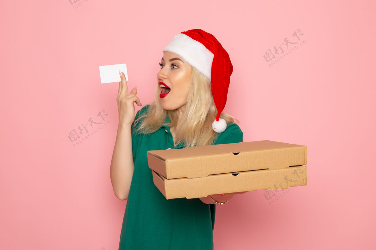 年轻的女性正面图：年轻女性手持银行卡和披萨盒 粉色墙面 彩色节日圣诞照片 工作服圣诞节披萨盒子