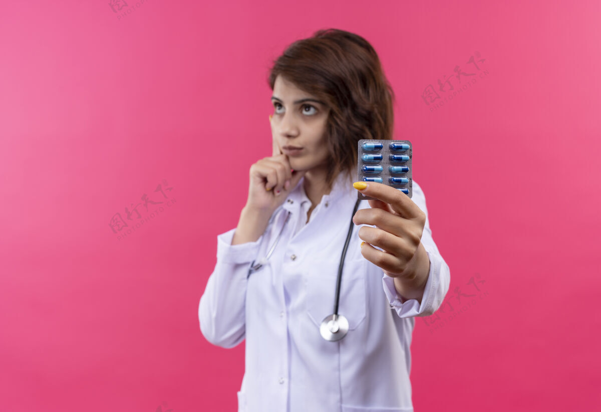 抱着一位年轻的女医生 穿着白大褂 手持听诊器 手里拿着水泡 手里拿着药丸 脸上带着沉思的表情 望着一旁听诊器外套看着