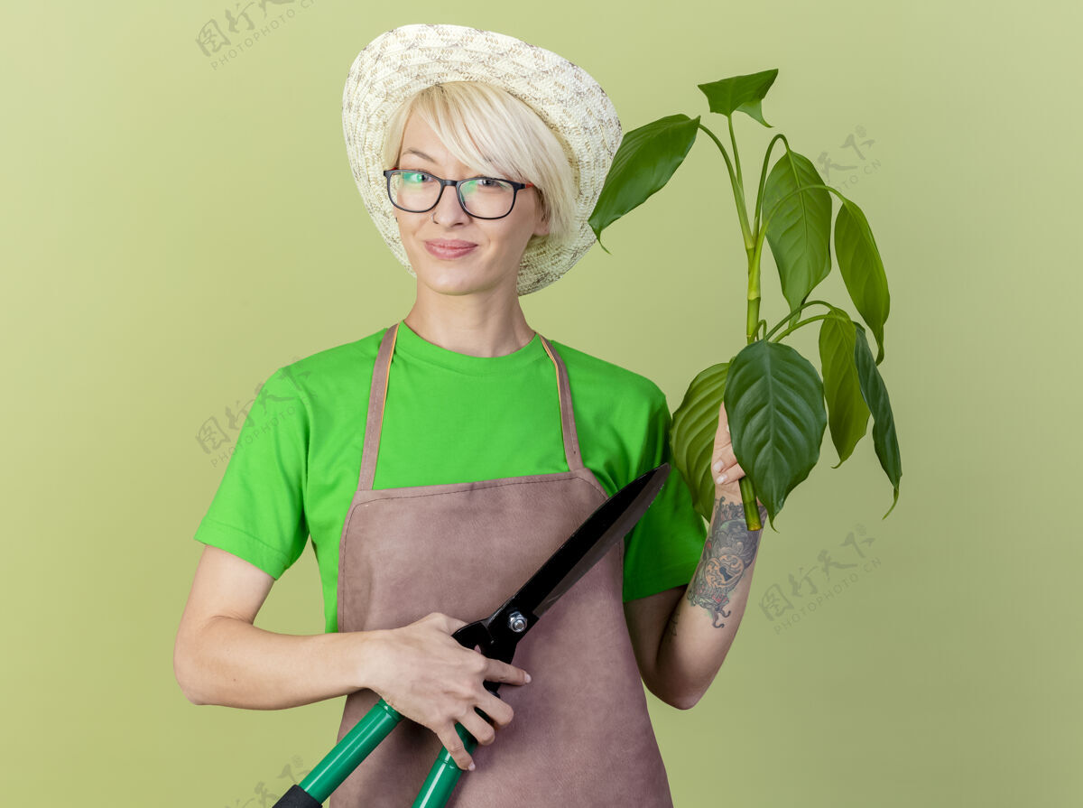 围裙一位年轻的园丁 围裙上留着短发 戴着帽子 手持植物和树篱剪 站在明亮的背景下 面带微笑地看着摄像机站着园丁女人