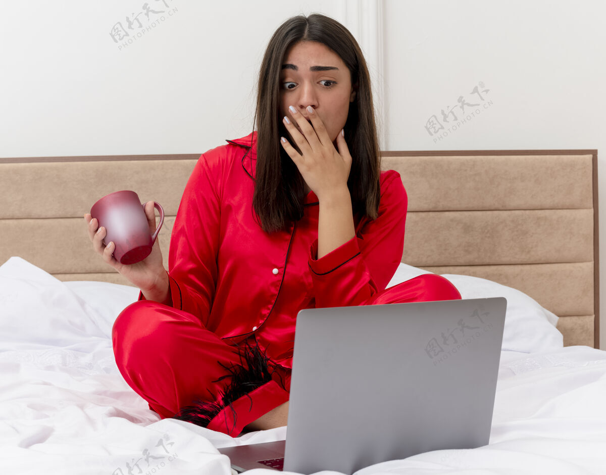惊讶穿着红色睡衣的年轻漂亮女人坐在床上 拿着笔记本电脑和一杯咖啡 在卧室内部的灯光背景下 看起来既惊讶又惊讶床漂亮咖啡