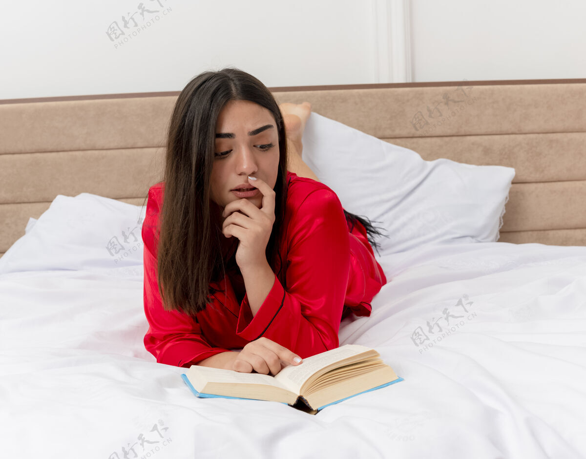 床穿着红色睡衣的年轻美女躺在床上看书 在卧室的室内灯光背景下显得很困惑红色年轻书