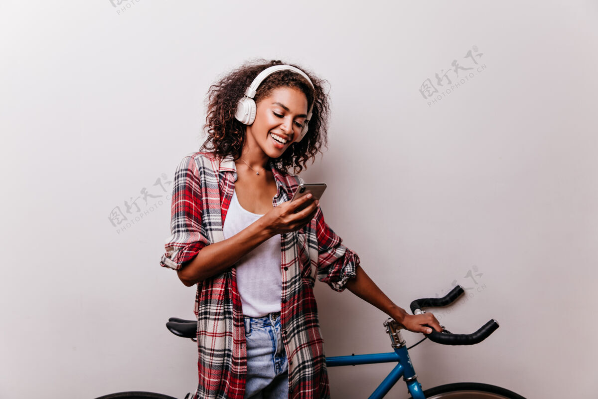 休闲戴着耳机看电话留言的迷人时尚女人穿着考究的非洲女孩拿着自行车拿着智能手机时尚休息活跃
