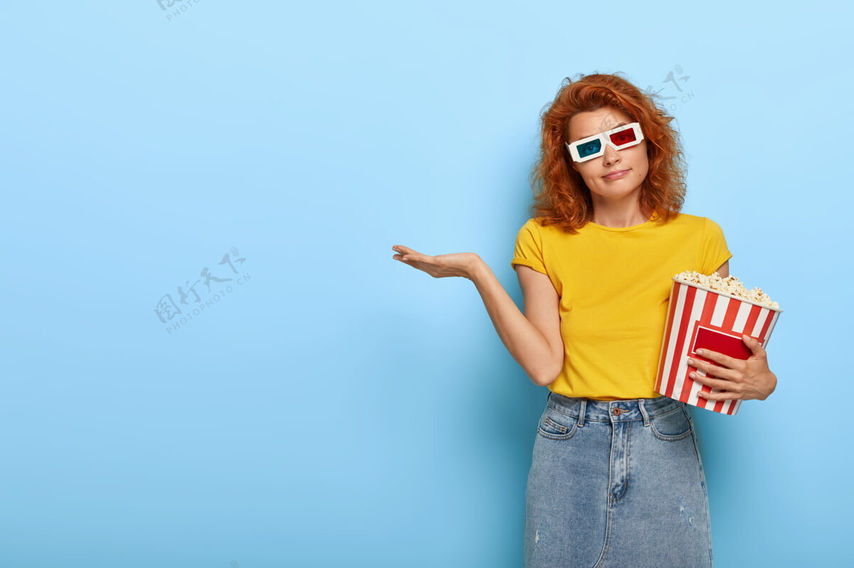 美味美丽的红发女人戴着虚拟眼镜 穿着黄色t恤和牛仔裙 抱着一篮子爆米花 走进电影院 表情疑惑 犹豫着看哪部电影空闲时间眼镜饮食