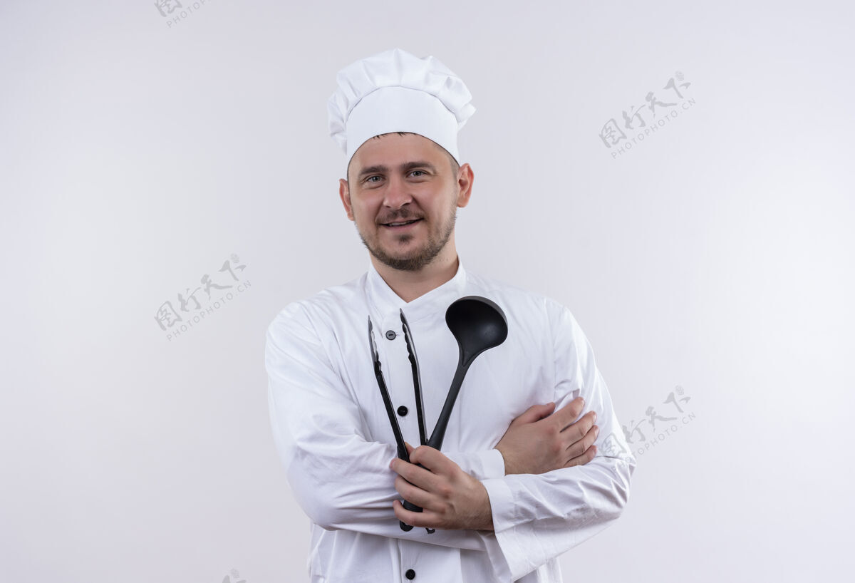 制服面带微笑的年轻帅哥厨师身着厨师制服 手里拿着勺子和烤架钳子 隔离在空白处厨师微笑年轻