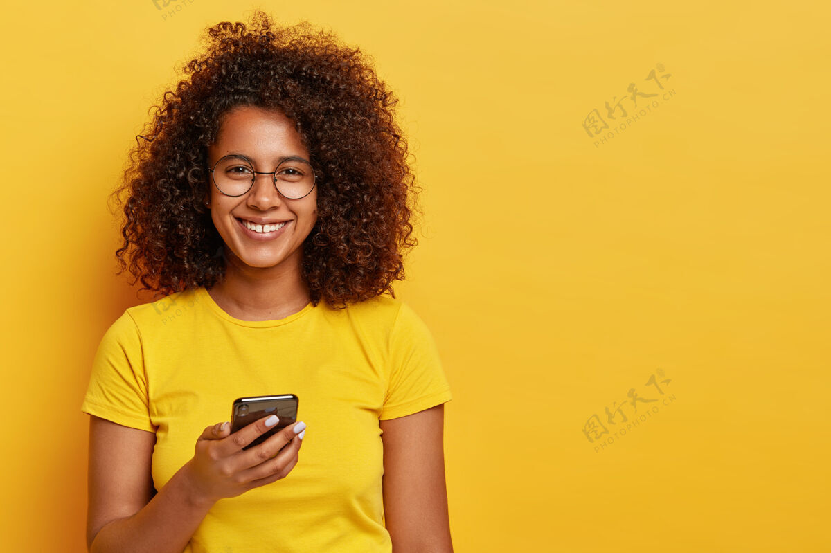 眼镜脸上带着愉快微笑的美女横拍 享受手机在线交流 阅读通知 戴着圆眼镜和黄色t恤科技和人的概念自由空间站使用