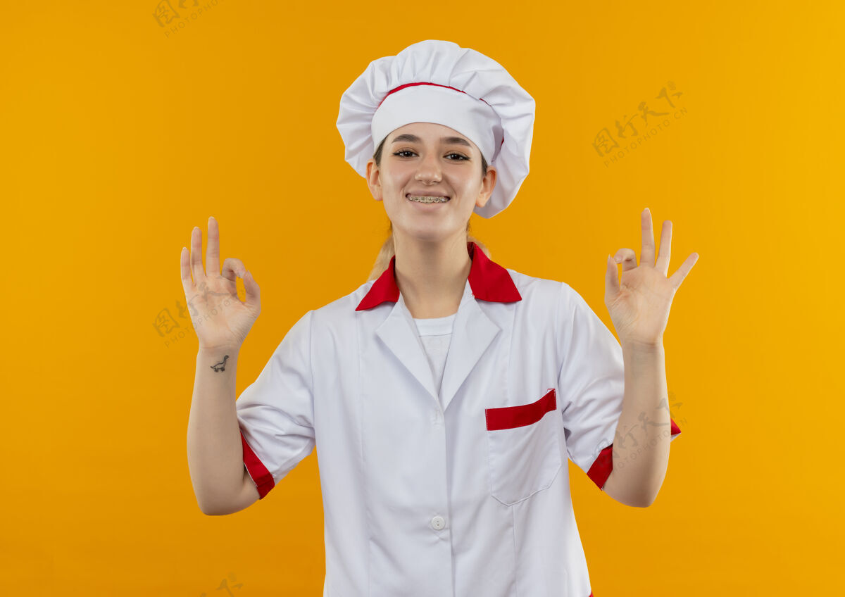 橙色微笑着的年轻漂亮厨师身着厨师制服 戴着牙套 在橙色的空间里做着“ok”的手势牙科好厨师