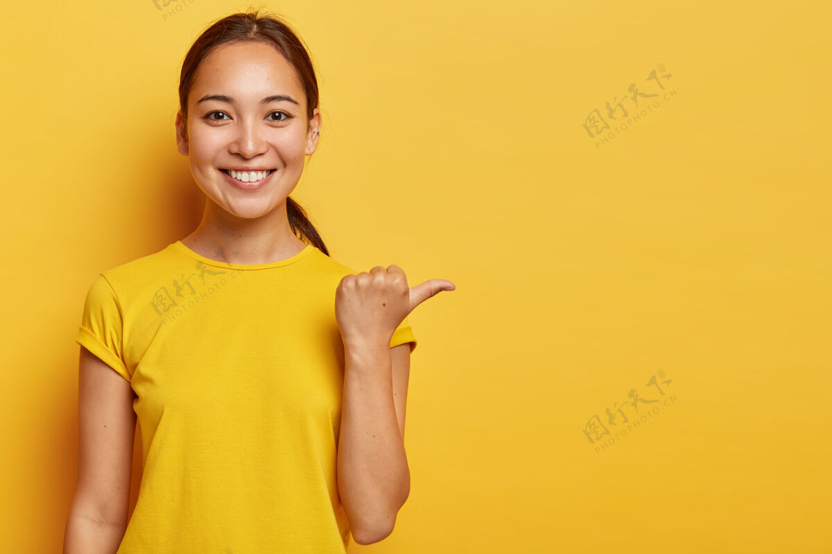 指尖年轻开朗的亚洲女性肖像用拇指指着远方 喜气洋洋的面部表情 为广告展示临摹空间 容貌宜人 穿着亮黄色的衣服年轻空外表