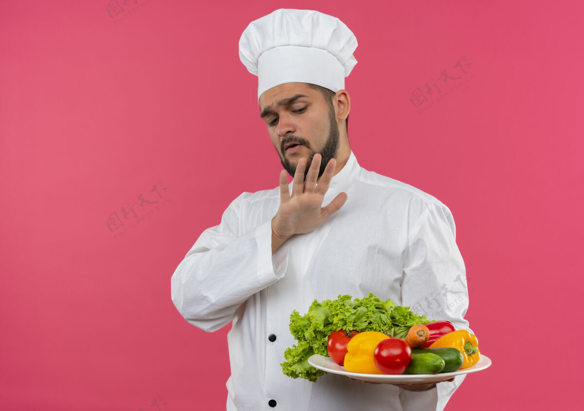 年轻未出狱的年轻男厨师身着厨师制服 手持并看着盘子里的蔬菜 在粉红色的空间里没有孤立的手势未发布蔬菜复制