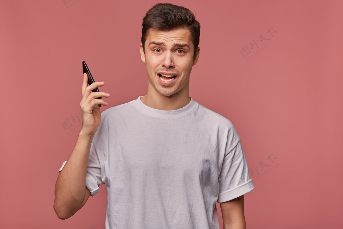 室内年轻惊讶的家伙穿着一件白t恤 拿着电话 听到难以置信的消息 表情激动地站在粉红色的地板上不可思议表情惊喜