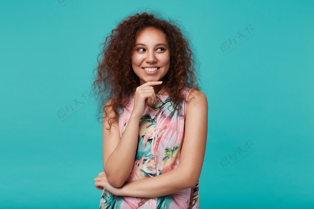 心情室内写真：快乐的年轻漂亮的卷发黑发女士抬起手托着下巴 面带迷人的微笑 孤零零地看着蓝色自然高兴黑发