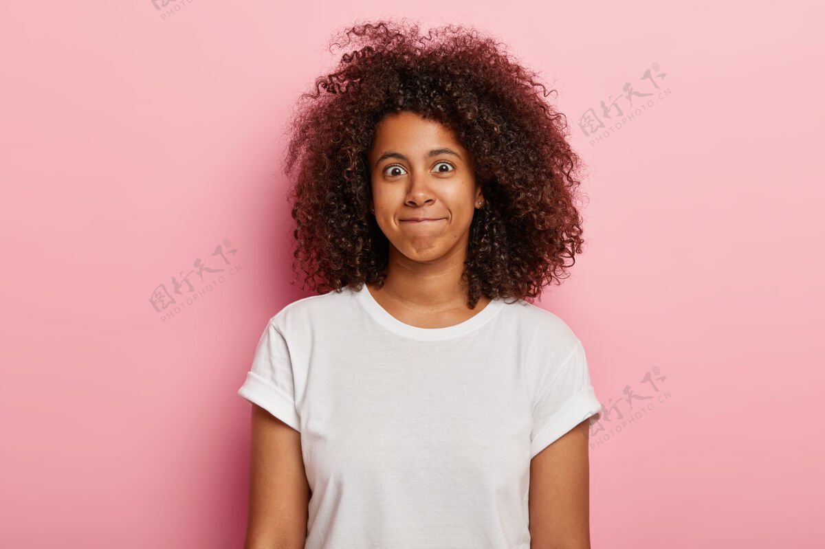 一瞥有趣女人的照片有卷曲浓密的头发 嘴唇贴在一起 有一张幸福的脸 穿着白色t恤 隔着粉红色的墙漂亮的年轻非洲裔美国女孩表达幸福看高兴乐观