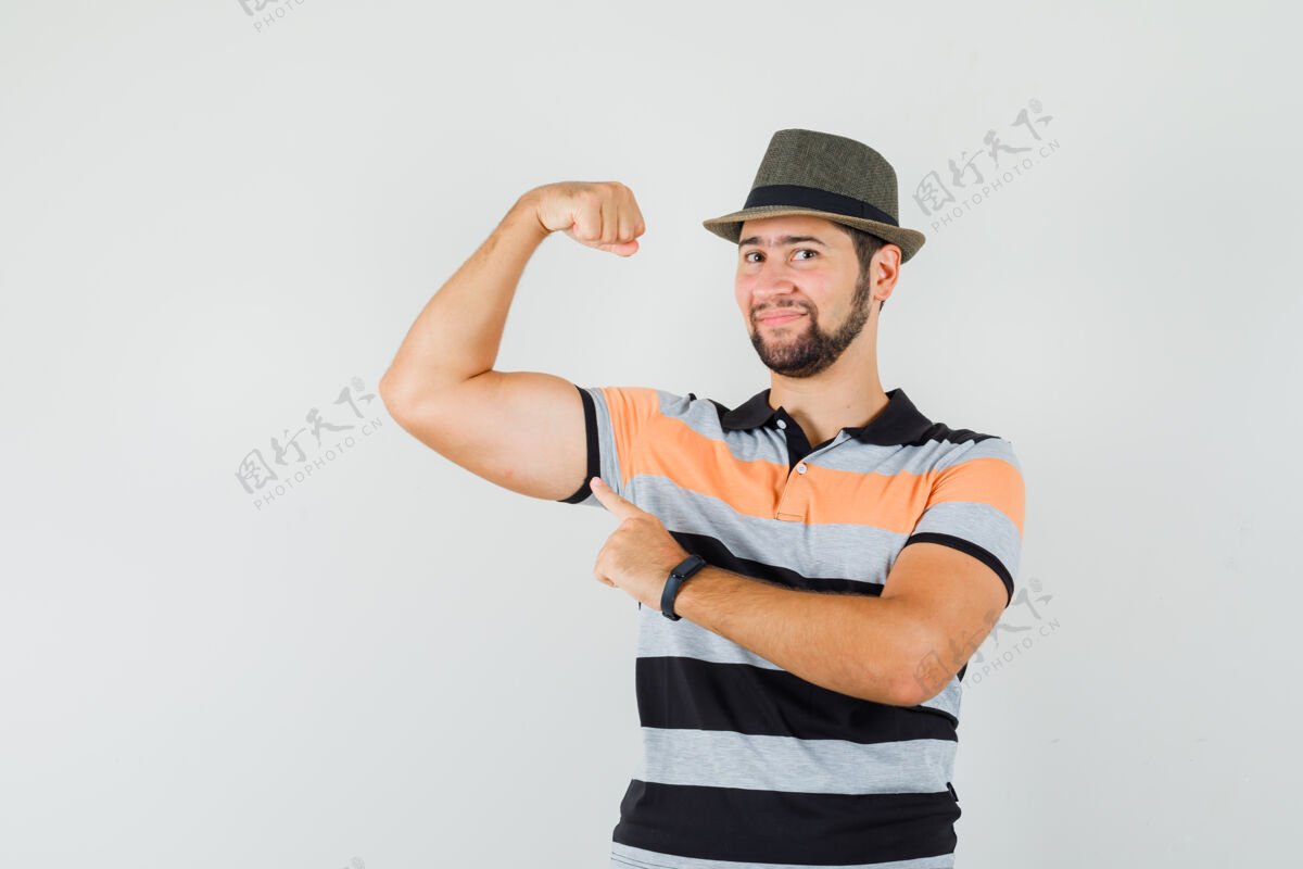 胡须年轻人指着他的肌肉 穿着t恤 戴着帽子 看起来很自信前视图友好帅气成功