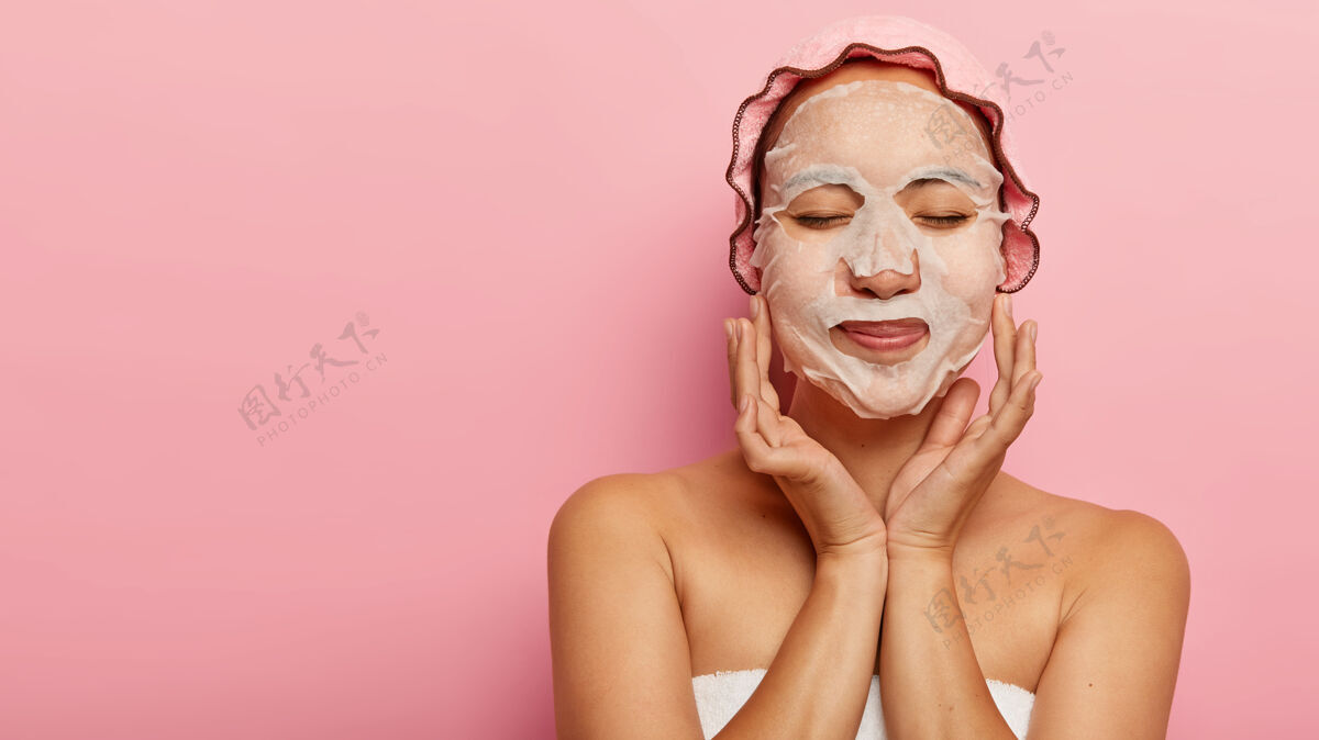 化妆品高兴的中国女人喜欢整容程序 有自然的纸面膜在脸颊上 裹在毛巾里 戴浴帽 闭着眼睛 隔离在粉红色的墙上 为您的广告腾出空间新鲜光滑乐观