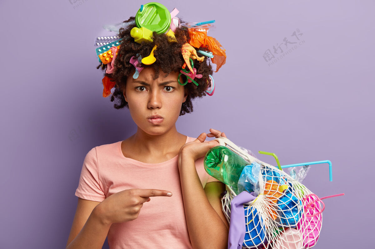 垃圾不高兴的非洲裔美国妇女对滥用塑料感到愤怒的照片 指着收集垃圾的袋子 头上有垃圾 隔离在紫色的墙上不可回收的污染概念关怀压力浪费