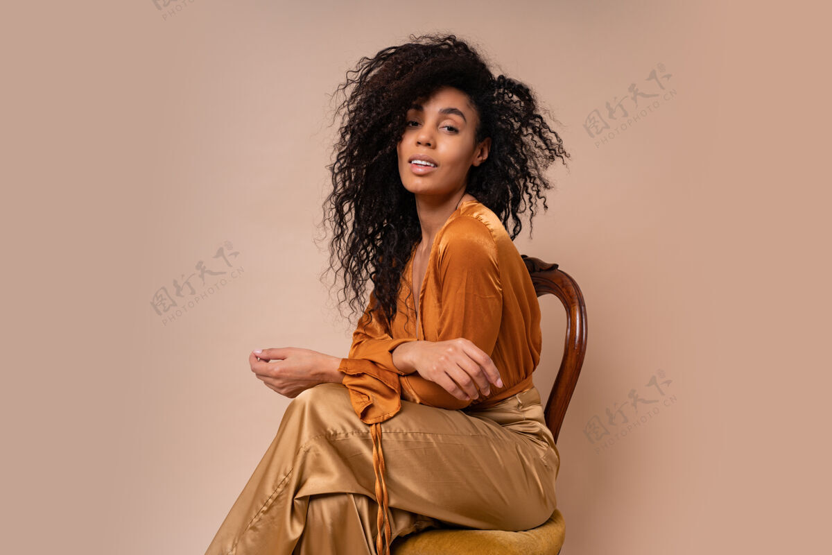 肖像时尚年轻诱人的非洲模特 完美的卷发 优雅的橙色衬衫和丝绸裤子 坐在老式椅子米色墙上模特时尚魅力