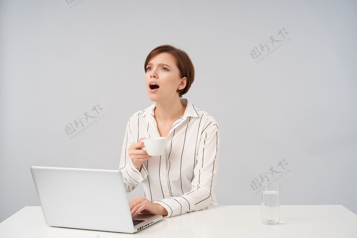 水年轻的黑发短发女人 随意的发型 一边看一边喊着什么 一边喝茶一边把手放在笔记本电脑的键盘上 孤零零地站在白色的地板上深色坐着办公室