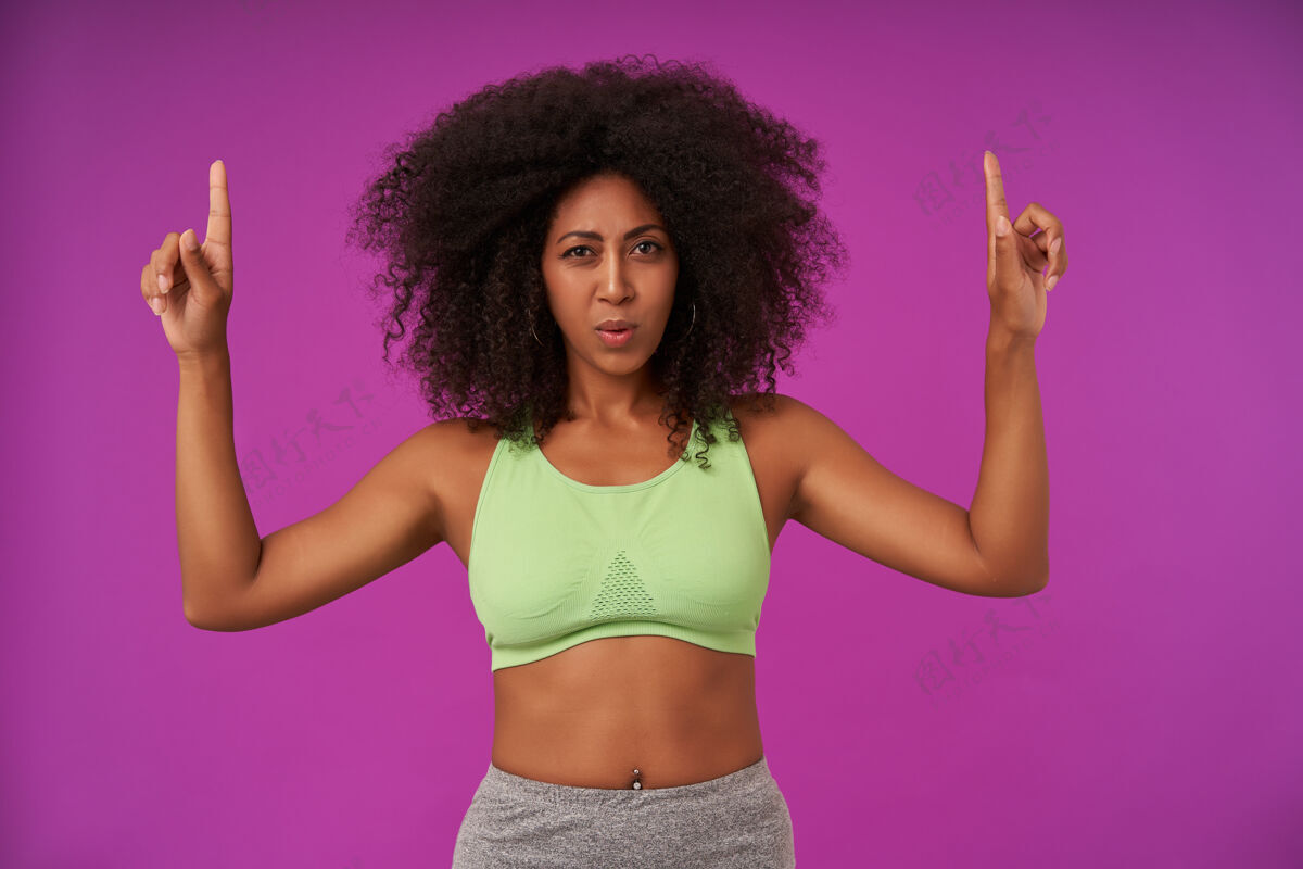 休闲年轻的黑皮肤运动女性 卷发女士 穿着浅绿色上衣 食指向上 斜视着 孤立在紫色上紫色指尖指数