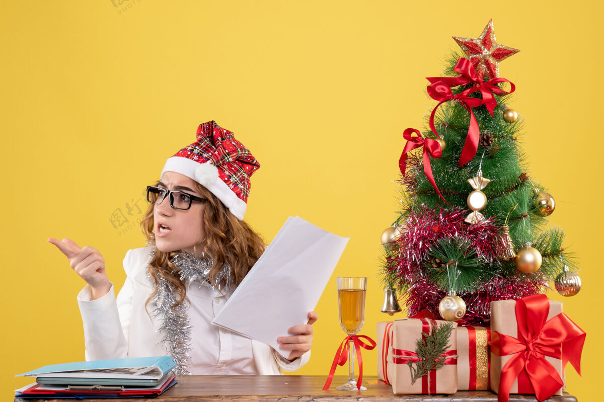 礼物前视图女医生坐在圣诞树上 拿着黄色背景上的文件医生圣诞树生活