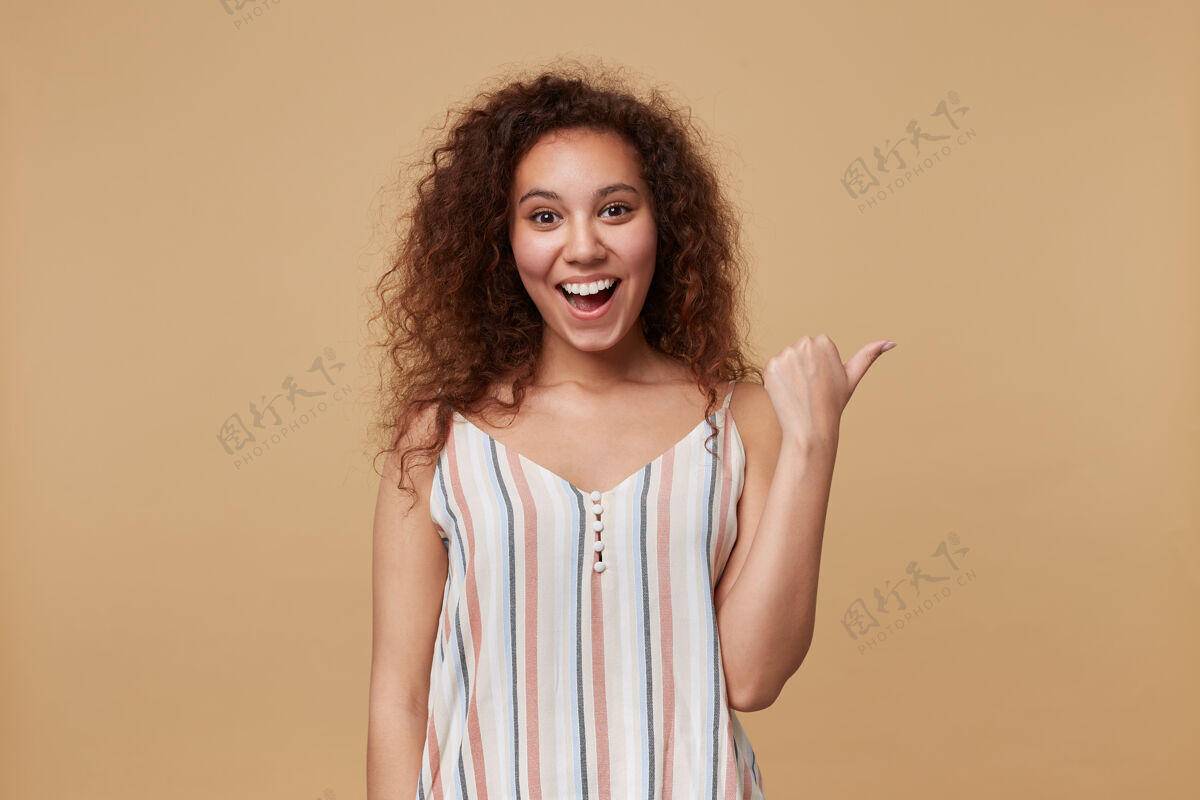 长年轻快乐的棕色头发卷曲的女性的画像快乐地笑 同时用拇指指向一边 在夏天的条纹上衣米色孤立室内提高年轻