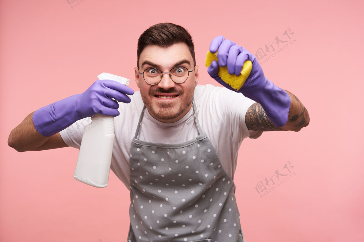深色一幅年轻的蓝眼睛黑发男性的画像 戴着眼镜 做鬼脸 手里拿着家用化学品 隔离在粉红色的地板上粉色休闲衣服