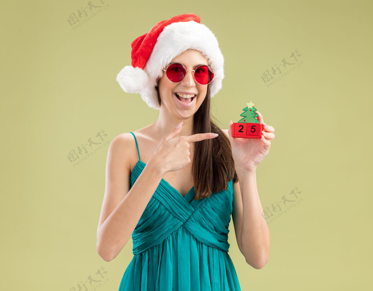 眼镜戴着太阳眼镜 戴着圣诞帽 指着圣诞树饰物的快乐白人女孩快乐装饰圣诞老人