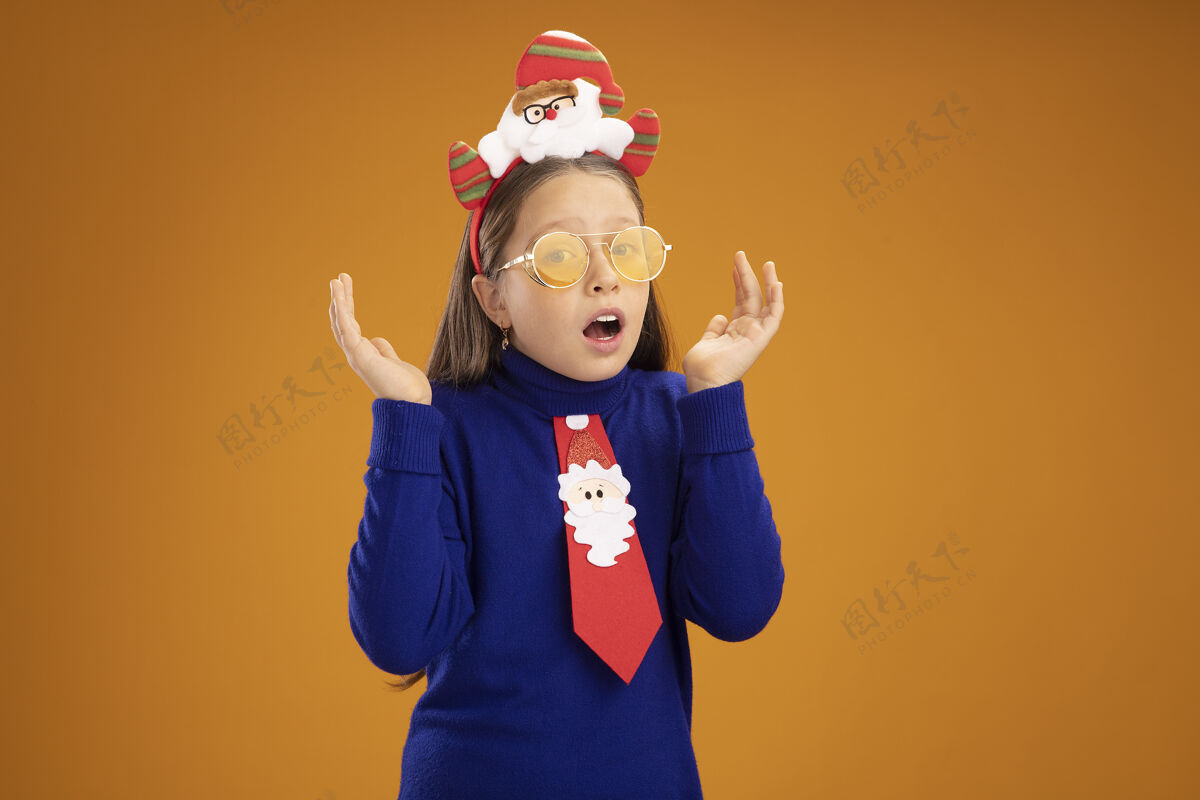 头小女孩穿着蓝色高领毛衣 打着红色领带 头上戴着有趣的圣诞戒指 困惑和惊讶地举起双臂红色领带边缘