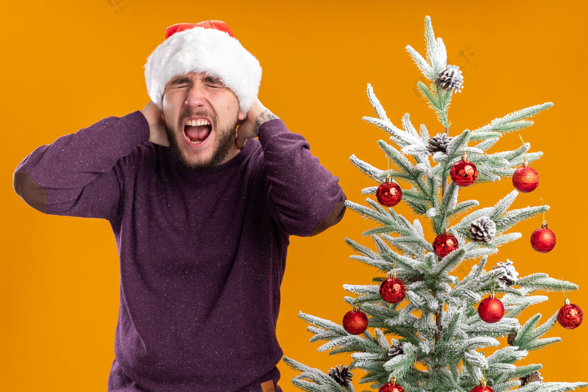 圣诞节身穿紫色毛衣 戴圣诞帽的年轻人站在橙色背景下的圣诞树旁 带着恼怒的表情大喊大叫表情站着帽子