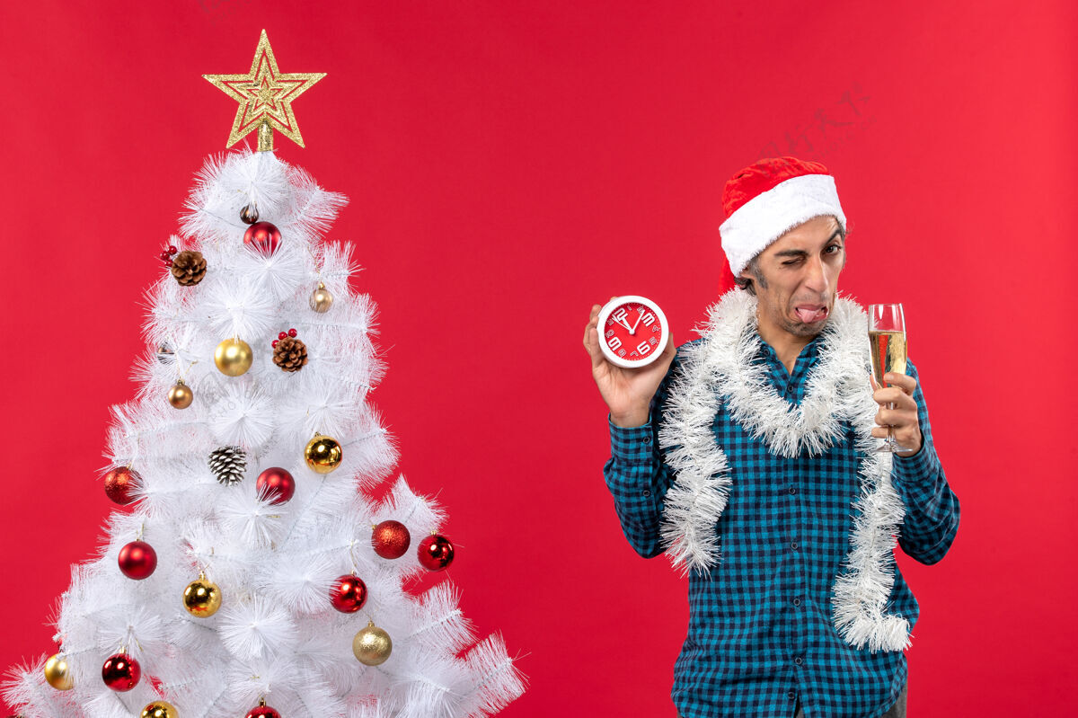 小伙子一个戴着圣诞老人帽子 拿着一杯葡萄酒和一个挂钟的酸溜溜的年轻人站在红色的圣诞树旁圣诞老人小伙子圣诞树