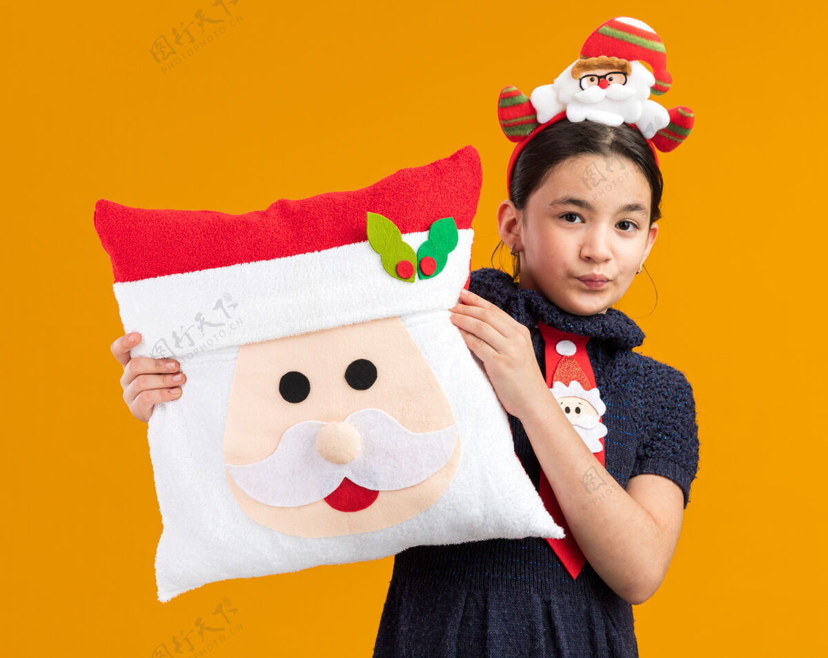 编织穿着针织连衣裙的快乐小女孩戴着红色领带 头上戴着滑稽的镶边 抱着圣诞枕头 面带微笑圣诞节新年脸