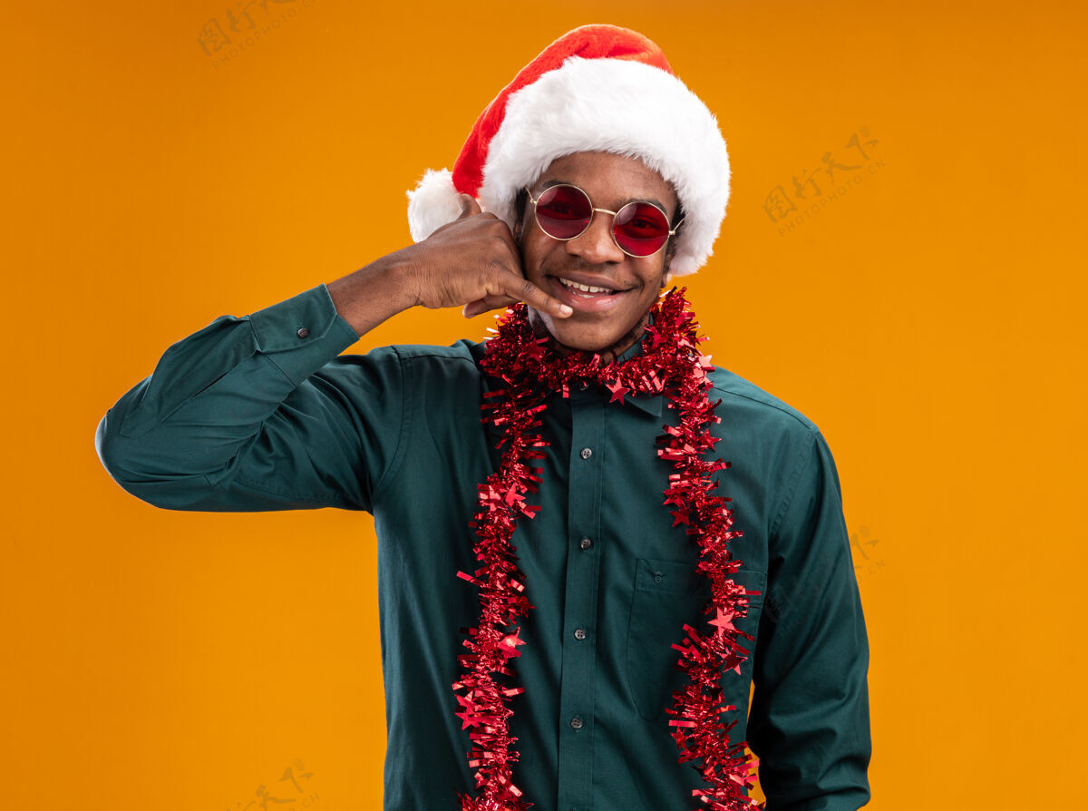 圣诞老人一个戴着圣诞帽 戴着花环 戴着墨镜的非裔美国人站在橘色的墙上愉快地微笑着做着“打电话给我”的手势愉快帽子制作