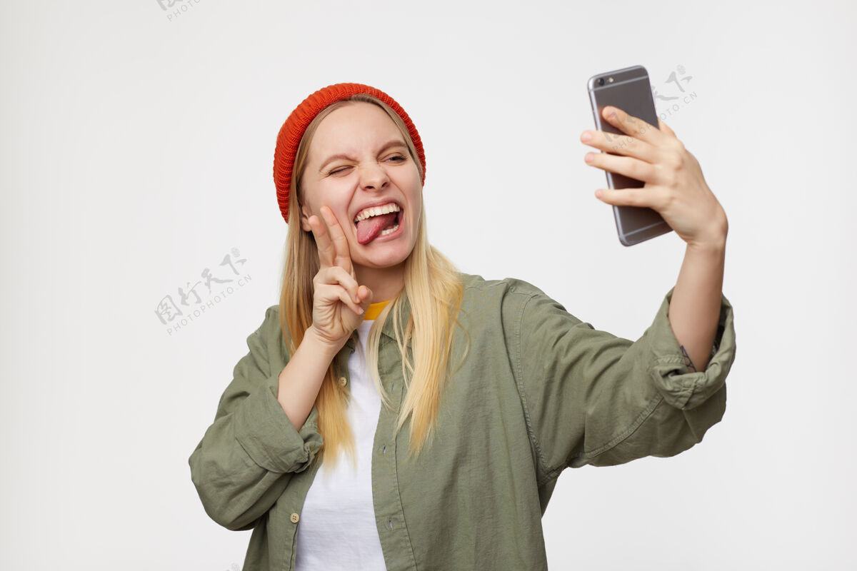 发型一张室内肖像 年轻漂亮的白头女士戴着红帽子 一边在智能手机上自拍一边做着滑稽的表情 表现出和平的姿态 在蓝色上与世隔绝年轻化妆愚弄