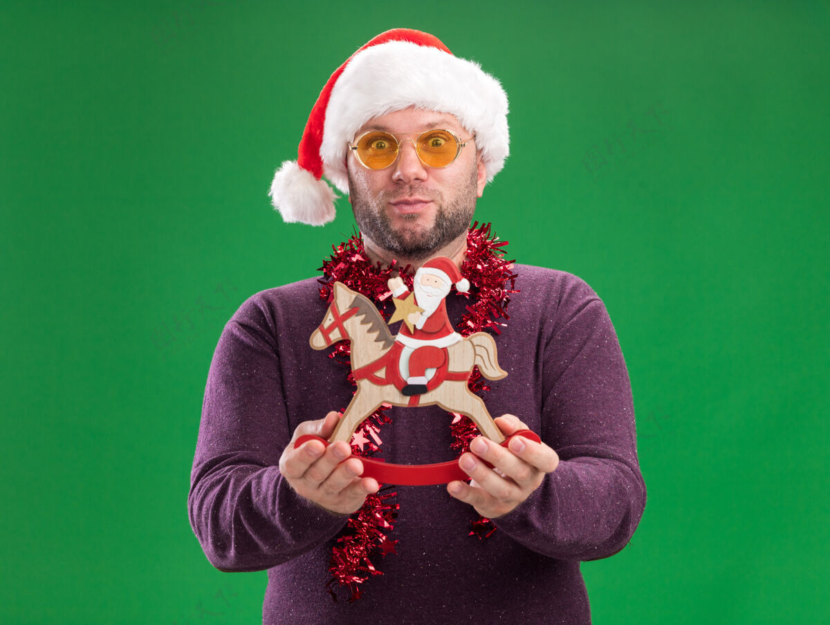 走向中年给人印象深刻的中年男子戴着圣诞帽 脖子上戴着金箔花环 戴着眼镜 圣诞老人站在绿色墙上的摇马雕像上花环圣诞老人金属丝
