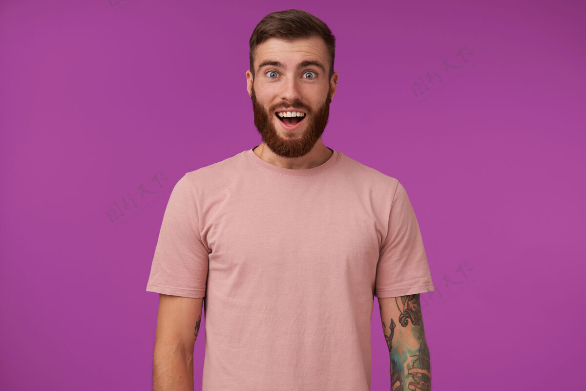 模特一个年轻的留着胡子的黑发男人的肖像 他身上有纹身 穿着米色t恤和时髦的配饰 站在紫色的地面上 双手放下 张大嘴巴米色衣服男性