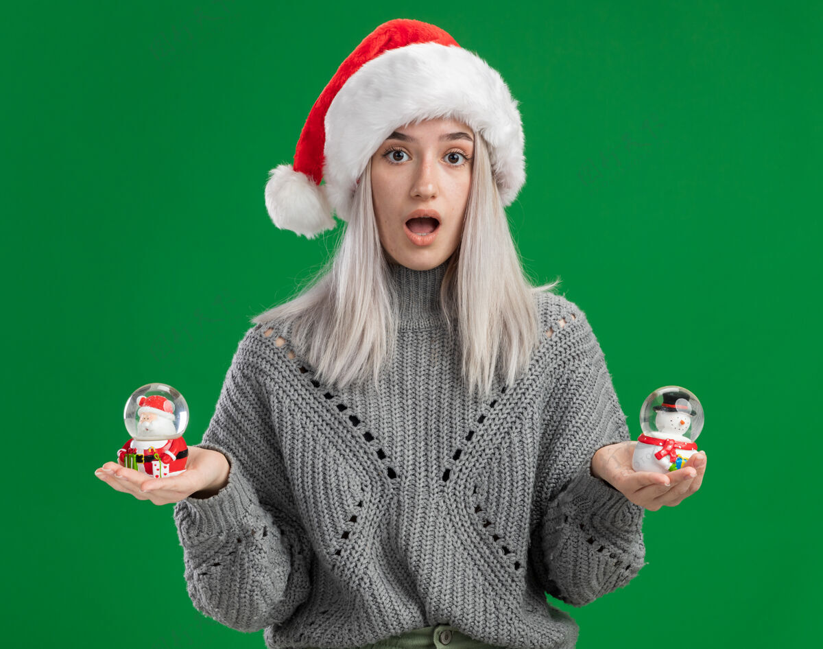 抱着年轻的金发女郎穿着冬天的毛衣 戴着圣诞帽 手里拿着圣诞玩具雪球 站在绿色的背景上惊讶地看着相机毛衣圣诞老人地球仪