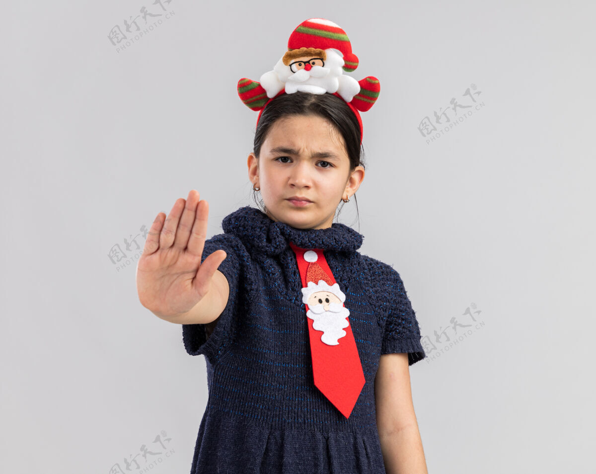 站着小女孩穿着针织连衣裙 头上系着一条红色领带 戴着滑稽的圣诞戒指 表情严肃 用手做着停止的手势小衣服领带