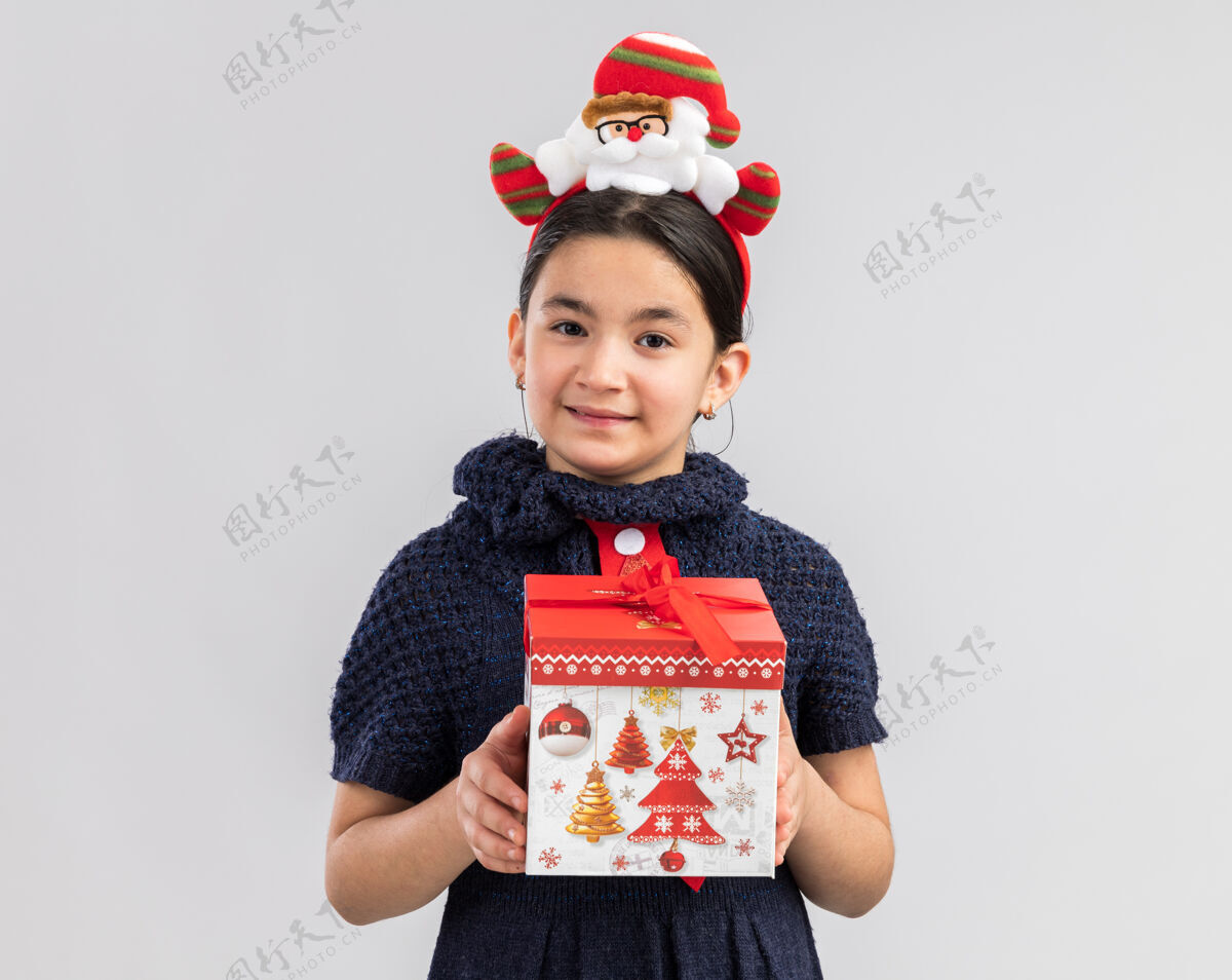 新小女孩穿着针织连衣裙 头上系着红色领带 头上戴着有趣的圣诞戒指 手里拿着圣诞礼物 脸上带着微笑 快乐而积极红色站着年