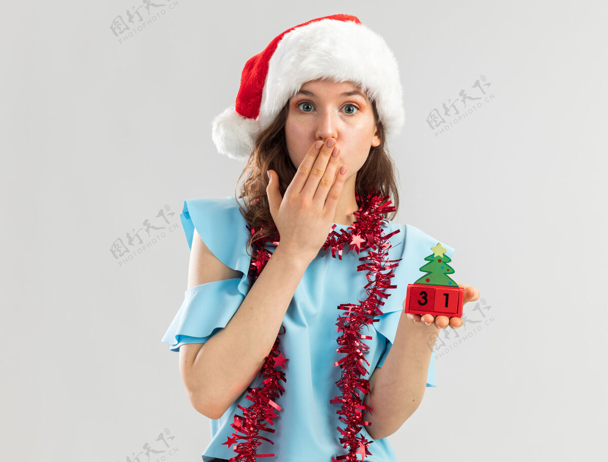 新年穿着蓝色上衣 戴着圣诞帽 脖子上戴着金属丝的年轻女子 手里拿着玩具立方体 看起来像是在用手捂住嘴巴嘴年被
