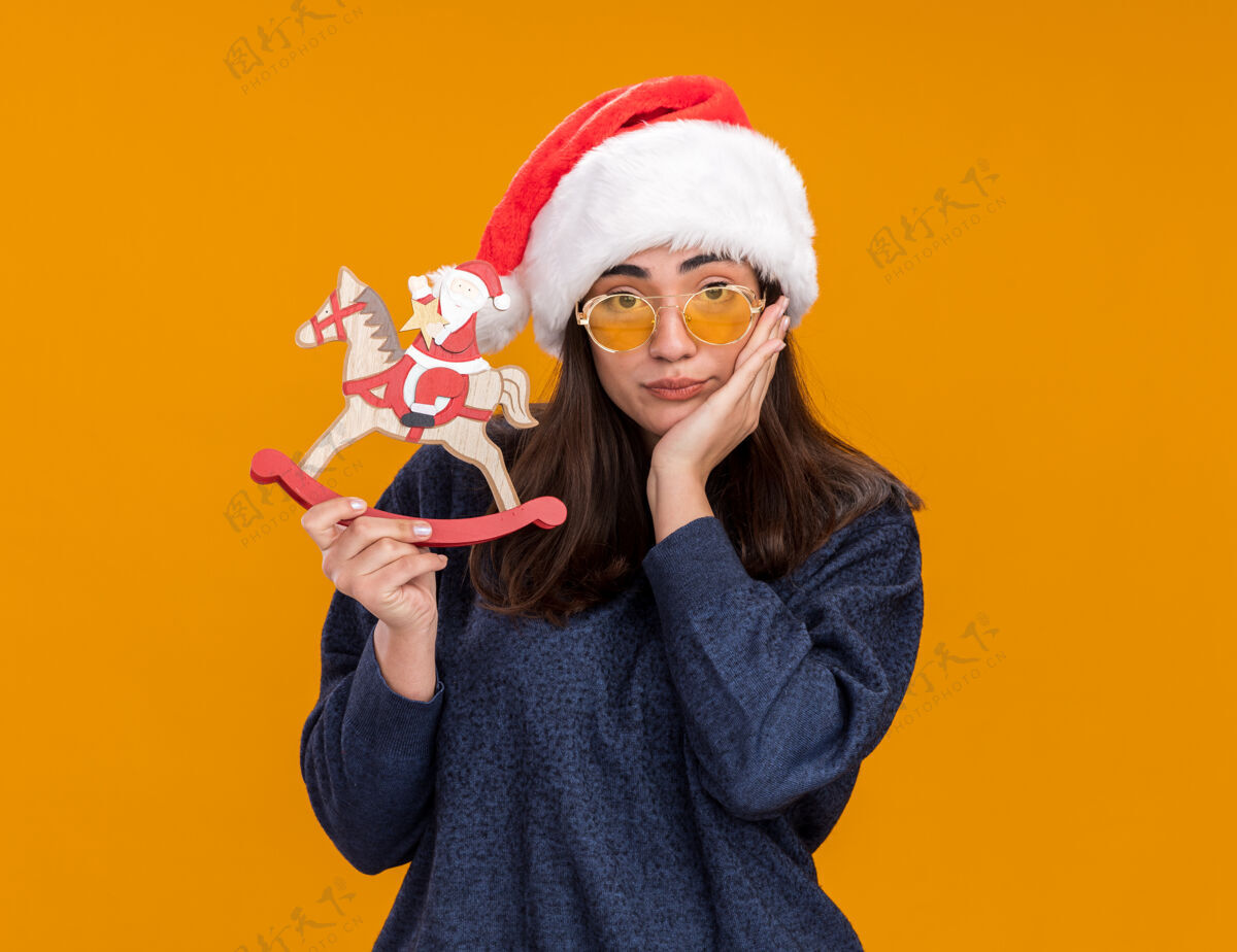 马戴着太阳眼镜 戴着圣诞帽的悲伤的白人女孩把手放在脸上 把圣诞老人抱在摇椅上脸装饰新年
