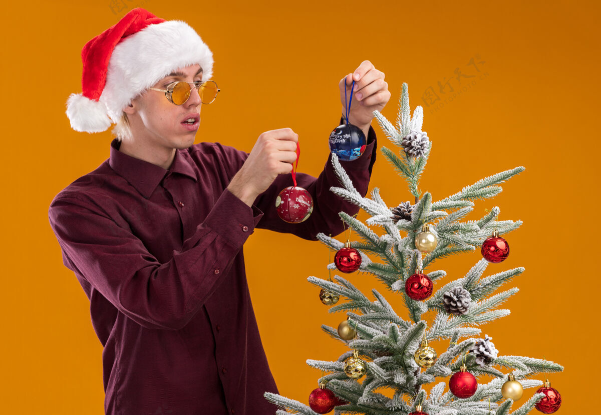 眼镜令人印象深刻的年轻金发男子戴着圣诞帽和眼镜站在侧面图装饰圣诞树附近的橙色背景穿着男人年轻