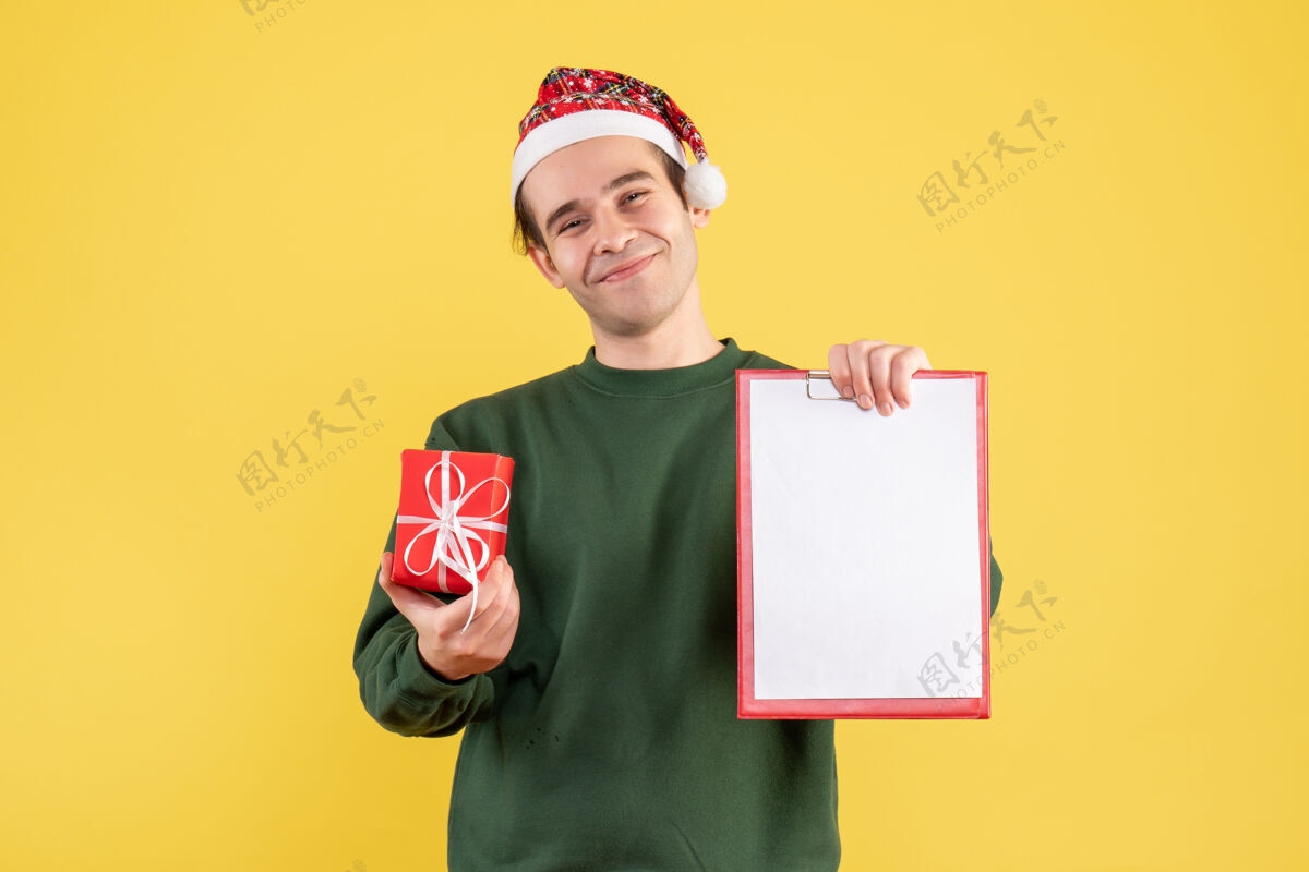 成人正面图：穿着绿色毛衣的年轻人拿着剪贴板和礼物站在黄色的地板上男人站着帅哥