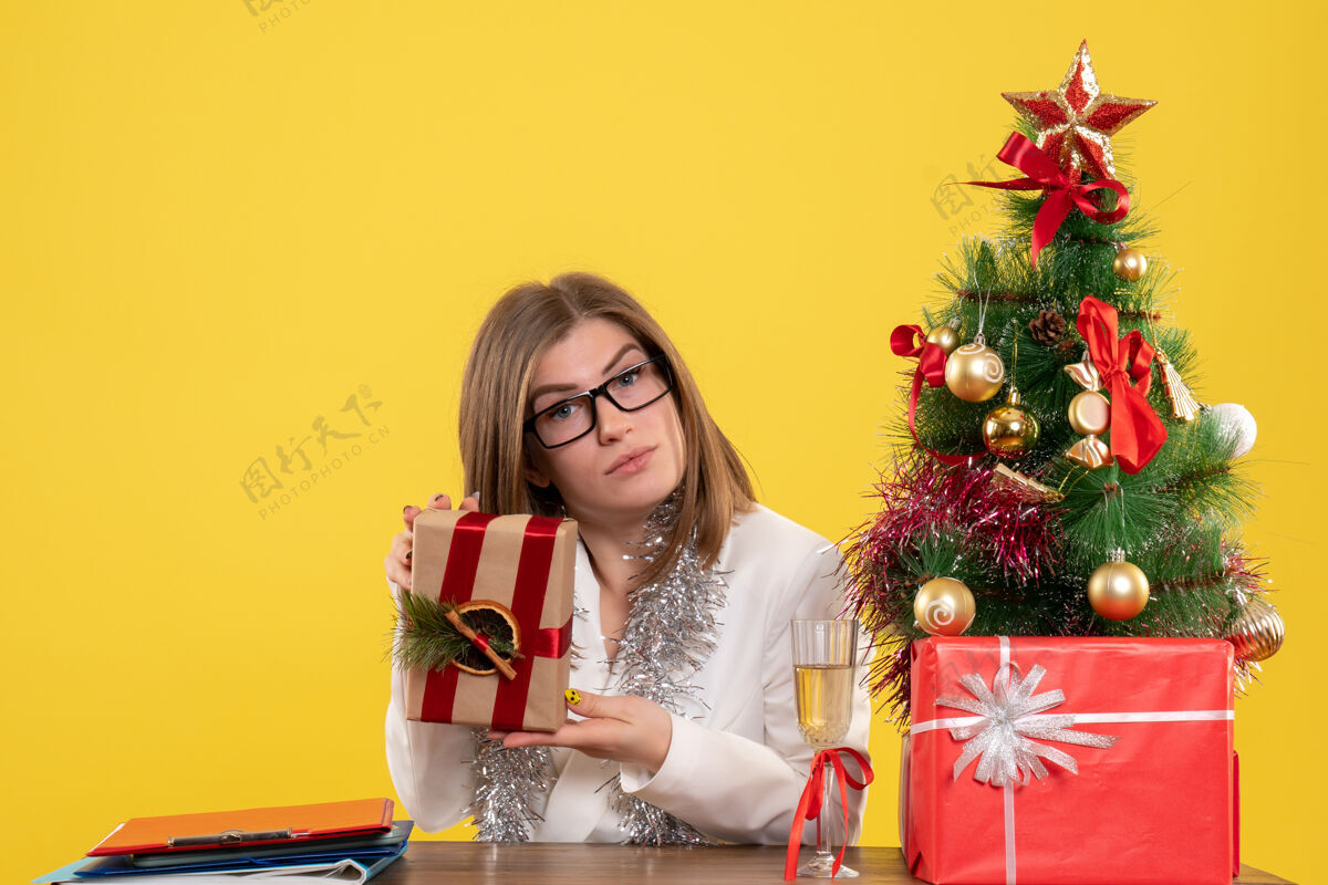 新正面图：女医生坐在桌子前 手里拿着黄色背景的礼物 还有圣诞树和礼品盒办公室花安排