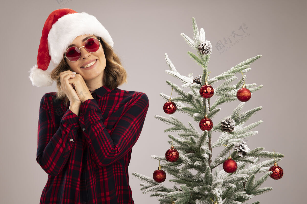 圣诞节高兴的年轻漂亮的女孩站在圣诞树旁边戴着圣诞帽和眼镜隔离在白色的墙上年轻戴着在附近
