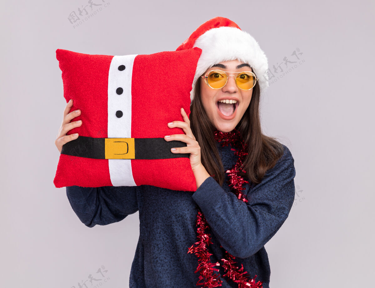 圣诞老人兴奋的年轻白人女孩戴着圣诞帽 脖子上戴着花环 拿着白色背景的装饰枕头 上面留有复制空间女孩新的圣诞快乐