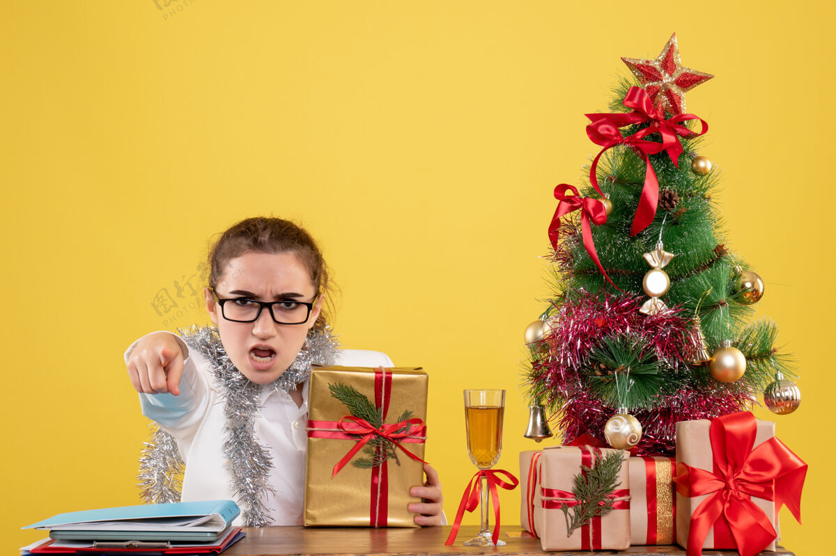 女性前视图：女医生坐在桌子后面 拿着圣诞礼物 黄色背景上的树花情感女医生