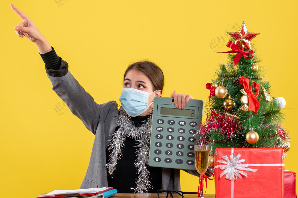 手指前视图好奇的女孩戴着医用面具坐在桌子旁拿着计算器手指指着圣诞树和礼物鸡尾酒坐着桌子好奇的