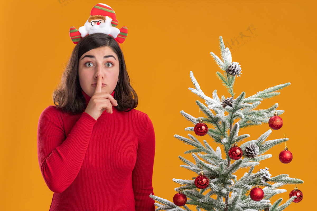 圣诞穿着圣诞毛衣的年轻女孩戴着滑稽的头带看着摄像机在橙色背景下的圣诞树旁做着沉默的手势毛衣头带穿着