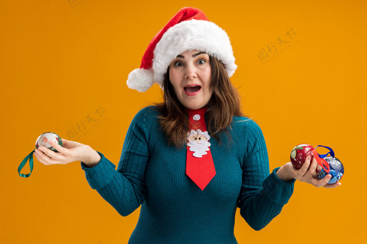 成人惊讶的成年白人妇女戴着圣诞帽 打着圣诞领带 手里拿着玻璃球饰品空间橙色领带